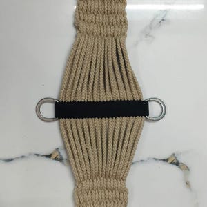 Puede incluir: Cincha de caballo de color beige con hebillas y anillas en D plateadas. La cincha está hecha de material tejido con una correa negra en el centro. Las hebillas tienen forma de D.