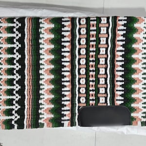 May include: A rectangular saddle pad with a woven design in dark green, white, and peach colors. The pad features vertical stripes and geometric patterns. A black leather item rests on the pad.