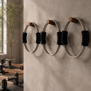 Puede incluir: Tres aros de Pilates montados en una pared texturizada. Cada aro es blanco con agarres acolchados negros y un soporte de madera. Los aros están espaciados uniformemente, lo que sugiere un estudio de fitness o gimnasio.