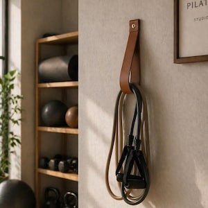 Op de afbeelding: Een bruine leren riem met een gouden klinknagel is aan een muur bevestigd en houdt twee weerstandsbanden vast. Eén band is beige, de andere is zwart. Op de achtergrond staat een houten plank met trainingsapparatuur.