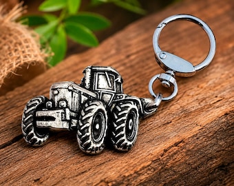 May include: A silver-toned metal keychain featuring a detailed tractor charm with prominent treaded tires, a cab, and a front grille. The tractor is attached to a circular split ring by a small swivel connector, creating a functional accessory.