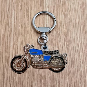 Puede incluir: Llavero de motocicleta plateado con depósito azul y asiento negro. El llavero tiene un anillo y un cierre plateados para sujetarlo a llaves o bolsos. El diseño de la motocicleta presenta ruedas y componentes del motor detallados.