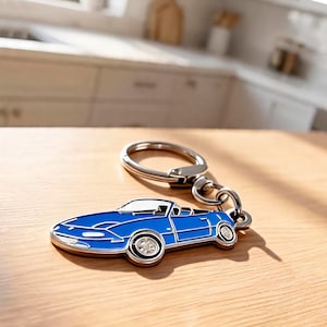 Peut inclure: Un porte-clés en émail bleu en forme de voiture de sport décapotable avec des contours en métal argenté et un anneau brisé circulaire. Le design de la voiture présente un profil bas, un pare-brise et deux roues avec des détails argentés.
