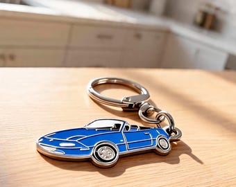 May include: A blue enamel keychain shaped like a convertible sports car with silver-toned metal outlines and a circular split ring attachment. The car design features a low profile, a windshield, and two wheels with silver-toned detailing.