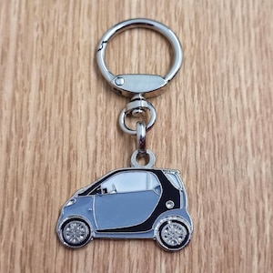 Op de afbeelding: Een zilveren sleutelhanger met een klein, grijs auto-scharnier. De auto heeft zwarte accenten en een wit interieur.