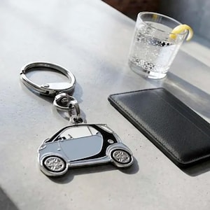 May include: A silver-toned metal keychain featuring a miniature car charm with a light blue body, black trim, and detailed wheels. The keychain includes a circular split ring and a swivel connector, presented alongside a black wallet and a glass.