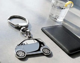 May include: A silver-toned metal keychain featuring a flat, stylized charm shaped like a small two-door hatchback car. The car charm has a light blue body, black trim, and detailed silver wheels. The keychain includes a circular split ring.