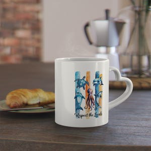 Könnte beinhalten: Weiße Keramik-Tasse mit herzförmigem Griff. Die Tasse zeigt ein Aquarelldesign von Meeresbewohnern, darunter eine Schildkröte, Delfine und einen Oktopus, mit dem Text "Respect the Locals". Das Design ist vor blauen und orangefarbenen vertikalen Streifen angeordnet.