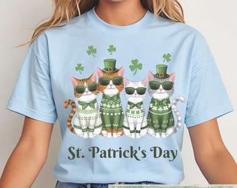 May include: A light blue t-shirt with a graphic of four cats wearing green sweaters and hats. The cats are wearing sunglasses and have green shamrocks around them. The text "St. Patrick's Day" is printed below the cats. PNG Digital Download Only is printed at the bottom of the shirt.