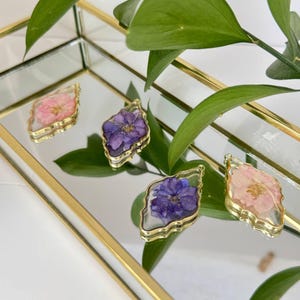 Puede incluir: Cuatro colgantes con marco dorado con flores secas en tonos rosa y morado. Los colgantes están colocados sobre una superficie espejada con ribete dorado, y hojas verdes en el fondo. Los colgantes tienen un revestimiento de resina transparente.