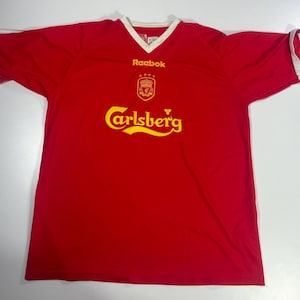 May include: Red short-sleeve jersey with a white V-neck collar and white sleeve cuffs. The jersey features the Reebok logo, a team crest, and the Carlsberg logo in yellow. The jersey is made of a lightweight material.