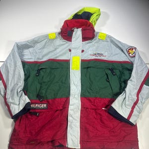 May include: A color-blocked Tommy Hilfiger sailing jacket featuring a light gray upper section, a dark green middle, and a red lower portion. It includes a high collar, a neon yellow hood, reflective accents, and various embroidered logos and text.