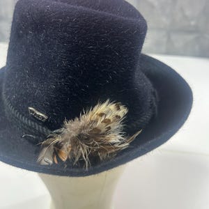 Könnte beinhalten: Ein dunkelmarineblauer Fedora-Hut mit einer dekorativen Feder und einem silberfarbenen Metallakzent. Der Hut hat eine strukturierte Krone und eine breite Krempe. Die Feder hat braune und cremefarbene Markierungen.