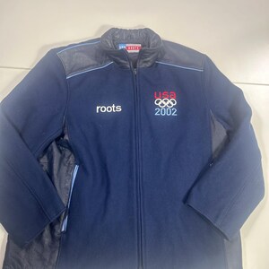 May include: Navy blue Roots zip-up jacket with leather accents. Features the USA Olympic logo and "2002" in red, white, and blue. The Roots logo is on the left chest. A classic, sporty outerwear piece.