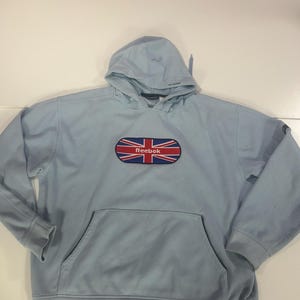 Op de afbeelding: Lichtblauwe hoodie met een kangoeroezak en een capuchon. De voorkant heeft een patch met de Union Jack-vlag en het Reebok-logo. De hoodie is gemaakt van een zacht materiaal en heeft lange mouwen.