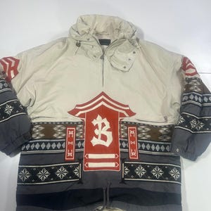 May include: Cream-coloured anorak jacket with a hood and a half-zip closure. The jacket features a large red and white emblem with the letter "B" on the front, along with patterned sleeves and lower section in shades of grey, brown, and black.