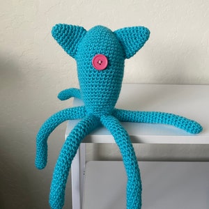 Op de afbeelding: Een handgemaakt turquoise gehaakt wezen met een knoop als oog en lange, tentakelachtige ledematen. Het pluche speelgoed heeft twee driehoekige oren en staat op een wit oppervlak. De knoop is roze.