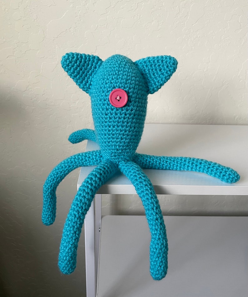 Op de afbeelding: Een handgemaakt turquoise gehaakt wezen met een knoop als oog en lange, tentakelachtige ledematen. Het pluche speelgoed heeft twee driehoekige oren en staat op een wit oppervlak. De knoop is roze.