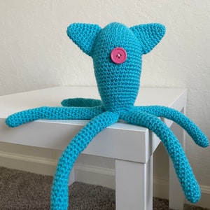 Op de afbeelding: Een handgemaakt turquoise gehaakt wezen met een knoopoog en vier lange tentakels. Het wezen heeft twee oorachtige uitsteeksels op zijn hoofd. Het speelgoed staat op een witte tafel.