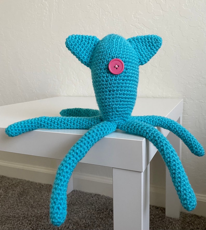 Op de afbeelding: Een handgemaakt turquoise gehaakt wezen met een knoopoog en vier lange tentakels. Het wezen heeft twee oorachtige uitsteeksels op zijn hoofd. Het speelgoed staat op een witte tafel.