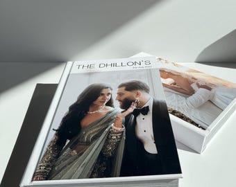 May include: A white hardcover photo album with the title "THE DHILLON'S" and the date "1st Aug 2022" printed on the front cover. The album features a photograph of a couple on the front cover. The woman is wearing a green and gold sari and the man is wearing a black tuxedo.
