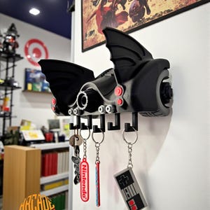 Pode incluir: Um suporte de chaves de parede preto em forma de veículo estilizado com asas de morcego, com detalhes circulares vermelhos e prateados. Possui cinco ganchos pretos pendurados na parte inferior, segurando três porta-chaves, incluindo um com o logótipo da Nintendo.