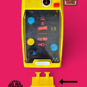 Op de afbeelding: Een geel handheld waterspel met een doorzichtige plastic kap, interne oranje en blauwe obstakels en een 'GOAL'-label. Onder het spel ligt een losse gele batterijklep, aangegeven door een zwarte pijl. De tekst luidt 'ONLY BATTERY COVER'.