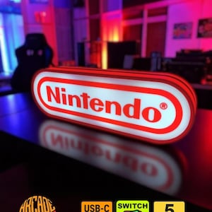 Può includere: Un'insegna rettangolare illuminata Nintendo con sfondo bianco, bordo rosso e testo rosso. L'insegna è dotata di una porta USB-C, un interruttore on/off e funziona a 5 volt. Il design include il logo Nintendo con il simbolo di marchio registrato.