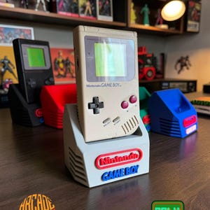Könnte beinhalten: Eine beige Nintendo Game Boy Handheld-Konsole in einem hellgrauen Kunststoff-Präsentationsständer. Der Ständer zeigt das rote Nintendo-Logo und den blauen Game Boy-Schriftzug auf der Vorderseite. Weitere Ständer in Schwarz, Rot, Grün und Blau sind sichtbar.