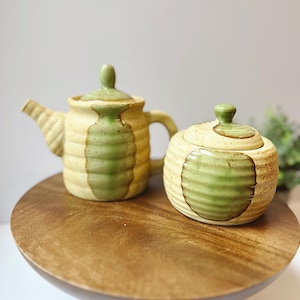 May include: A ceramic teapot and sugar bowl set. The teapot has a spout and handle, with a lid. The sugar bowl has a lid. Both pieces are beige with green horizontal stripes and sit on a wooden cake stand.