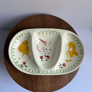 May include: Oval, three-sectioned ceramic serving dish with a white base. The dish features a hand-painted design of a bunny and two chicks. Green trim and red flowers decorate the dish, which is placed on a wooden surface.