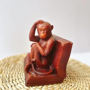 May include: A reddish-brown monkey figurine seated on a book-shaped base. The monkey is posed with one hand on its head, appearing thoughtful. The figurine is made of a smooth material and sits on a woven mat.