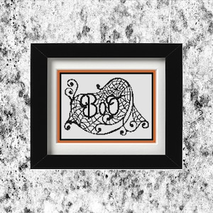 May include: A framed cross-stitch artwork featuring the word "Boo" in a spiderweb design. The black frame surrounds a white background with an orange border. A Halloween-themed decoration.