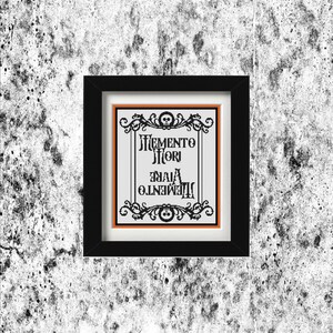 Könnte beinhalten: Ein gerahmtes Kunstwerk mit den Worten "Memento Mori, Vive Memento" in einer gotischen Schriftart. Das Kunstwerk befindet sich in einem schwarzen Rahmen mit einem orangefarbenen Rand und Totenkopfakzenten, vor einer strukturierten grau-weißen Wand.