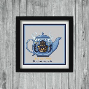 May include: A framed cross-stitch artwork featuring a blue teapot with intricate designs, including a Hamsa hand and an eye symbol. The teapot has a decorative lid and spout. The text "It is the Here, Do what thou wilt" is below the teapot.