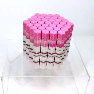 May include: A geometric sculpture composed of numerous hexagonal columns. The columns are arranged in a cube shape, with a gradient of colors from pink to white and brown. The sculpture is displayed on a clear acrylic surface.