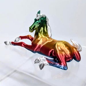May include: A colorful, rainbow-hued horse figurine in a reclining pose. The sculpture features a gradient of colors, including red, orange, yellow, green, and blue. The mane and tail are white. The horse is resting on a clear surface.