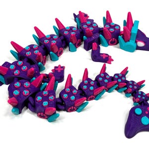 May include: A purple articulated dragon toy with pink and teal accents. The dragon's body is made up of segments with button details, and it has a matching game controller at the end. The toy is designed to resemble a video game theme.
