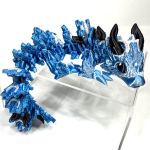 May include: A blue and white articulated dragon figurine. The dragon has a detailed, crystalline design with black horns and ears. The dragon is posed on a clear surface.