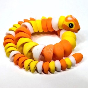 May include: A colorful, articulated snake toy in shades of orange, yellow, and white. The toy is designed with a segmented body, allowing for flexible movement. The snake's head features black eyes and a detailed, rounded shape. The toy is a fun and engaging item.