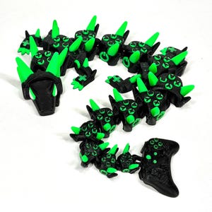 May include: A collection of black and green 3D-printed articulated dragon figures. Each dragon segment features green spikes and multiple green eye-like details. The figures are arranged on a white surface.
