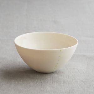 Może przedstawiać: Kremowa ceramiczna miska o zaokrąglonym kształcie. Seria małych, jasnozielonych kropek ułożona jest pionowo wzdłuż boku miski. Miska stoi na neutralnym szarym tle.