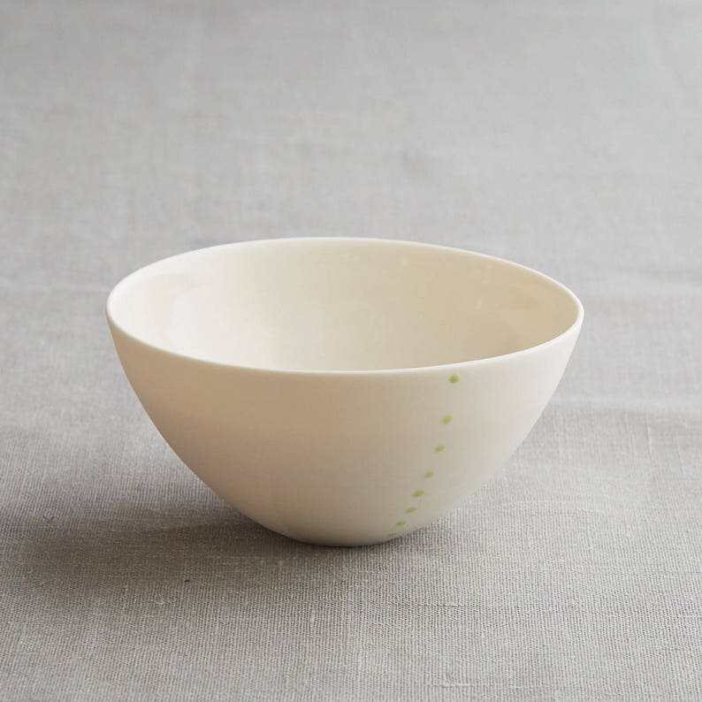 Może przedstawiać: Kremowa ceramiczna miska o zaokrąglonym kształcie. Seria małych, jasnozielonych kropek ułożona jest pionowo wzdłuż boku miski. Miska stoi na neutralnym szarym tle.