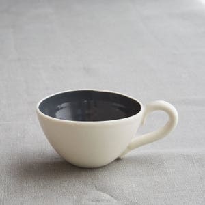 May include: A white ceramic teacup with a dark gray interior and a curved handle. The cup has a smooth, glossy finish and sits on a gray fabric surface. The design is simple and modern.