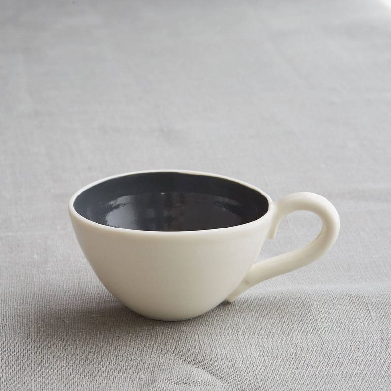 May include: A white ceramic teacup with a dark gray interior and a curved handle. The cup has a smooth, glossy finish and sits on a gray fabric surface. The design is simple and modern.
