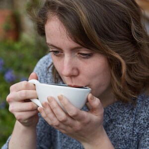 May include: A person drinking from a white ceramic coffee cup with a dark interior. The cup has a curved handle and is held with both hands. The person is wearing a gray sweater, and the background is blurred with greenery.