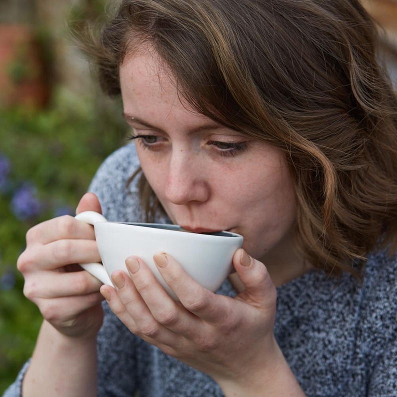 May include: A person drinking from a white ceramic coffee cup with a dark interior. The cup has a curved handle and is held with both hands. The person is wearing a gray sweater, and the background is blurred with greenery.