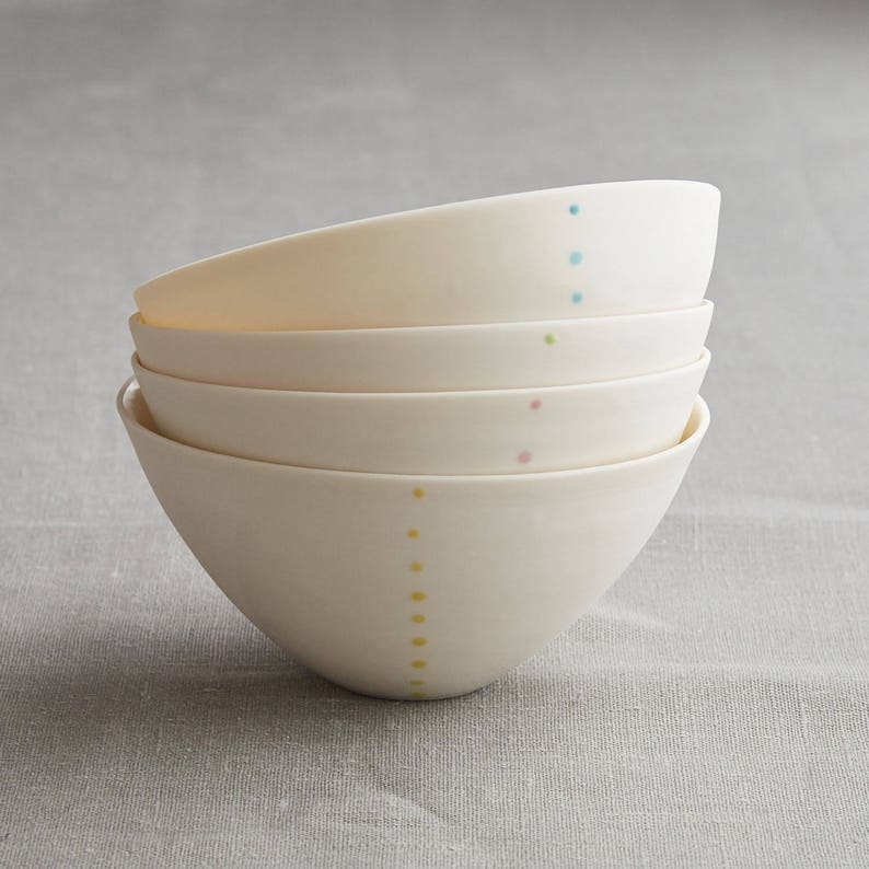 Może przedstawiać: Stos czterech kremowych ceramicznych misek. Każda miska ma szereg małych, kolorowych kropek w pionowej linii. Kropki są w odcieniach ż&oacute;łtego, r&oacute;żowego, zielonego i niebieskiego. Miski są ułożone jedna na drugiej.