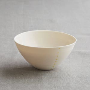 Może przedstawiać: Kremowa ceramiczna miska o prostym, nowoczesnym designie. Miska ma gładkie, matowe wykończenie i lekko asymetryczny kształt. Seria małych, ż&oacute;łtych kropek jest ułożona pionowo wzdłuż boku miski.
