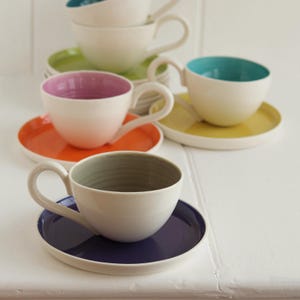 May include: A collection of white ceramic teacups and saucers in various colors. The cups have colored interiors and are paired with matching or complementary colored saucers. The colors include orange, yellow, green, purple, and blue.